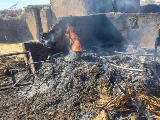 In Al Faschir sieht man auf diesem Foto von einem Haus nur noch brennende Trümmer und Teile der Grundmauern, die noch ca. einen Meter hoch stehen. Eine Person in einer weißen Hose schaut auf die kohlenden Trümmer.