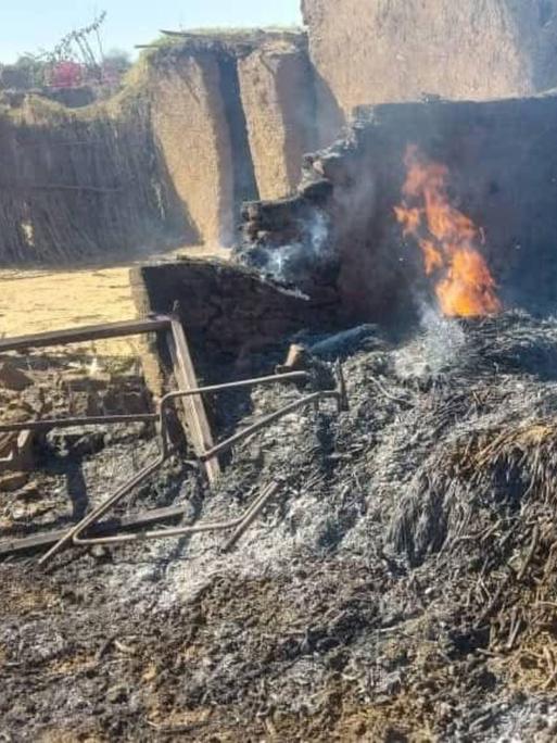 In Al Faschir sieht man auf diesem Foto von einem Haus nur noch brennende Trümmer und Teile der Grundmauern, die noch ca. einen Meter hoch stehen. Eine Person in einer weißen Hose schaut auf die kohlenden Trümmer.