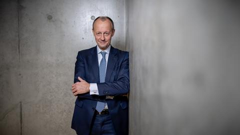 Friedrich Merz steht in einem dunkelblauen Anzug, einer blau gemusterten Krawatte vor einer Wand aus Sichtbeton. Mit verschränkte Armen schaut er den Betracht direkt an.