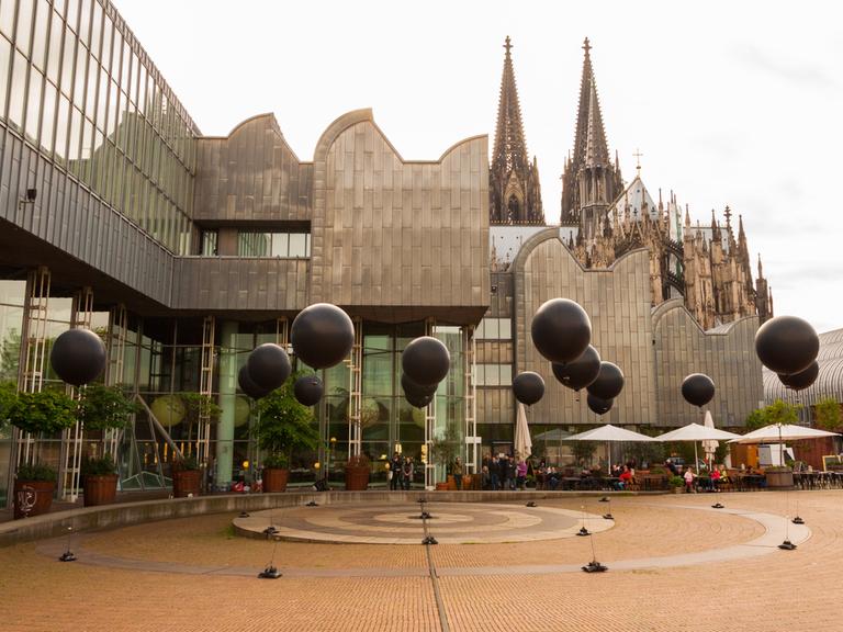 Vor der Kulisse von Philharmonie und Kölner Dom schweben sechzehn schwarze Ballons in der Luft, die eine musikalische Installation  darstellen.