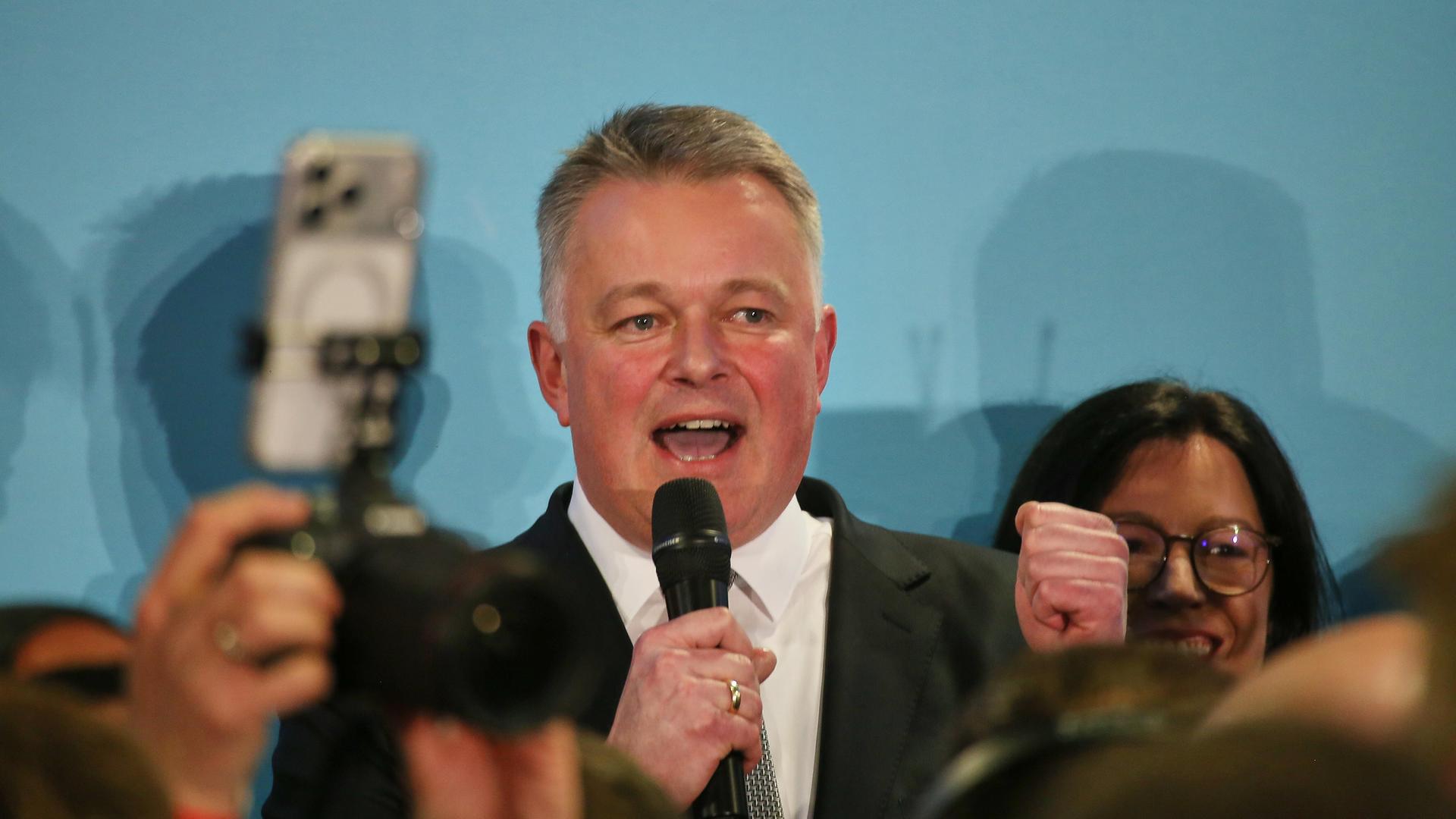 Gordon Schnieder steht vor einer türkisfarbenen Wand und spricht in ein Mikrofon. Er ballt die linke Faust und lächelt. Gordon Schnieder steht vor einer türkisfarbenen Wand und spricht in ein Mikrofon. Er ballt die linke Faust und lächelt.