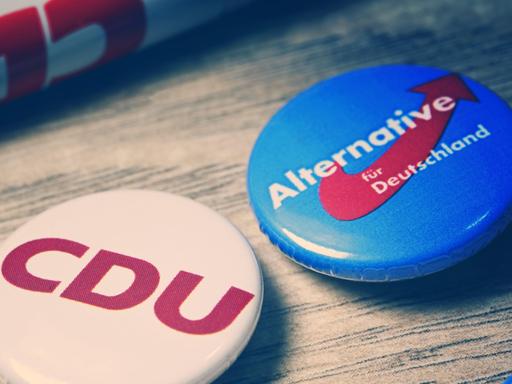 Partei-Anstecker mit Logos von CDU und AfD liegen auf einer Holzplatte