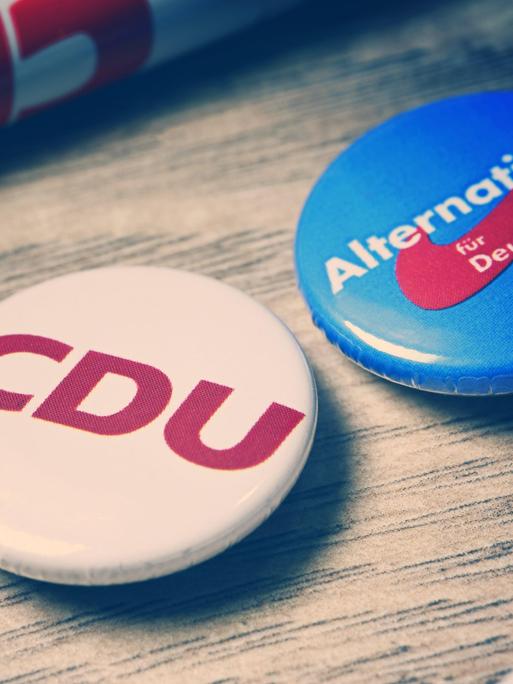 Partei-Anstecker mit Logos von CDU und AfD liegen auf einer Holzplatte