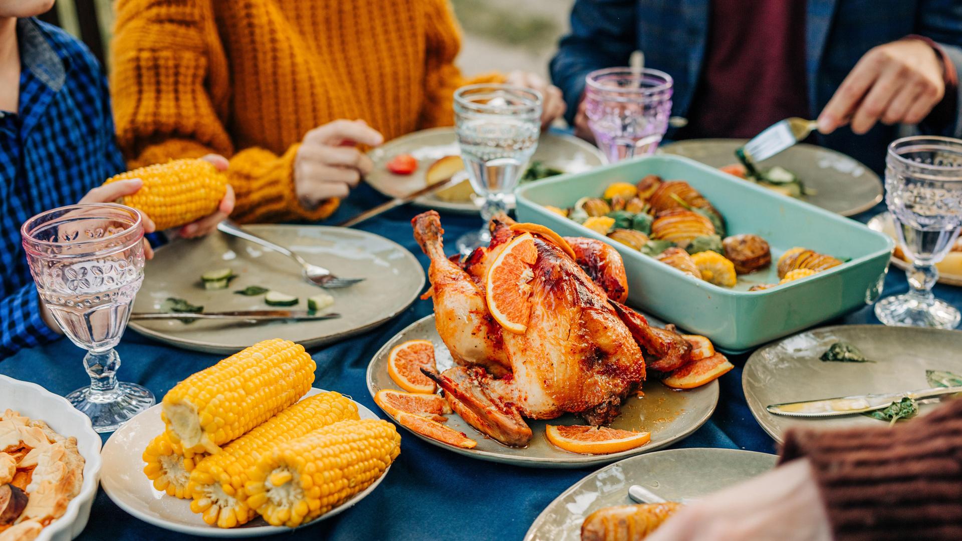 An einem reich gedecktem Tisch mit Maiskolben, Geflügelbraten und Geschirr sitzen mehrere Personen und essn An einem reich gedecktem Tisch mit Maiskolben, Geflügelbraten und Geschirr sitzen mehrere Personen und essn
