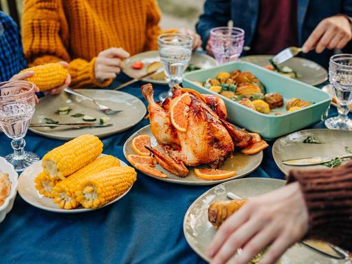 An einem reich gedecktem Tisch mit Maiskolben, Geflügelbraten und Geschirr sitzen mehrere Personen und essn