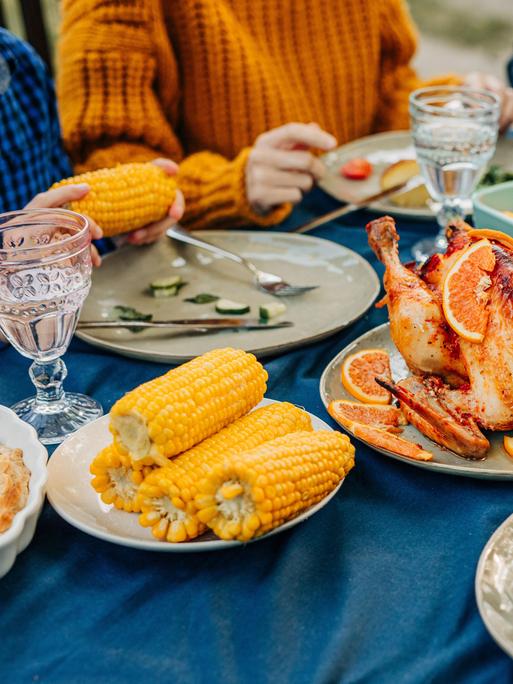 An einem reich gedecktem Tisch mit Maiskolben, Geflügelbraten und Geschirr sitzen mehrere Personen und essn