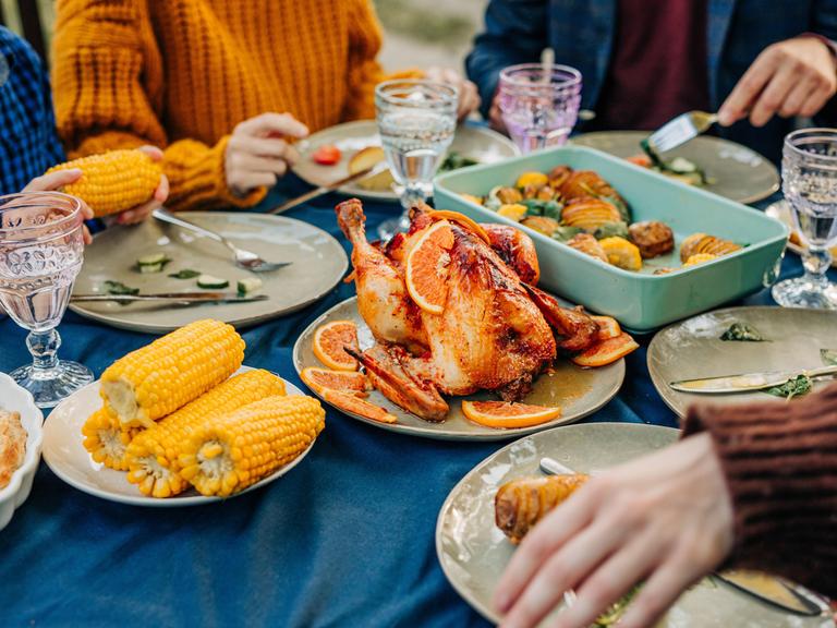 An einem reich gedecktem Tisch mit Maiskolben, Geflügelbraten und Geschirr sitzen mehrere Personen und essn