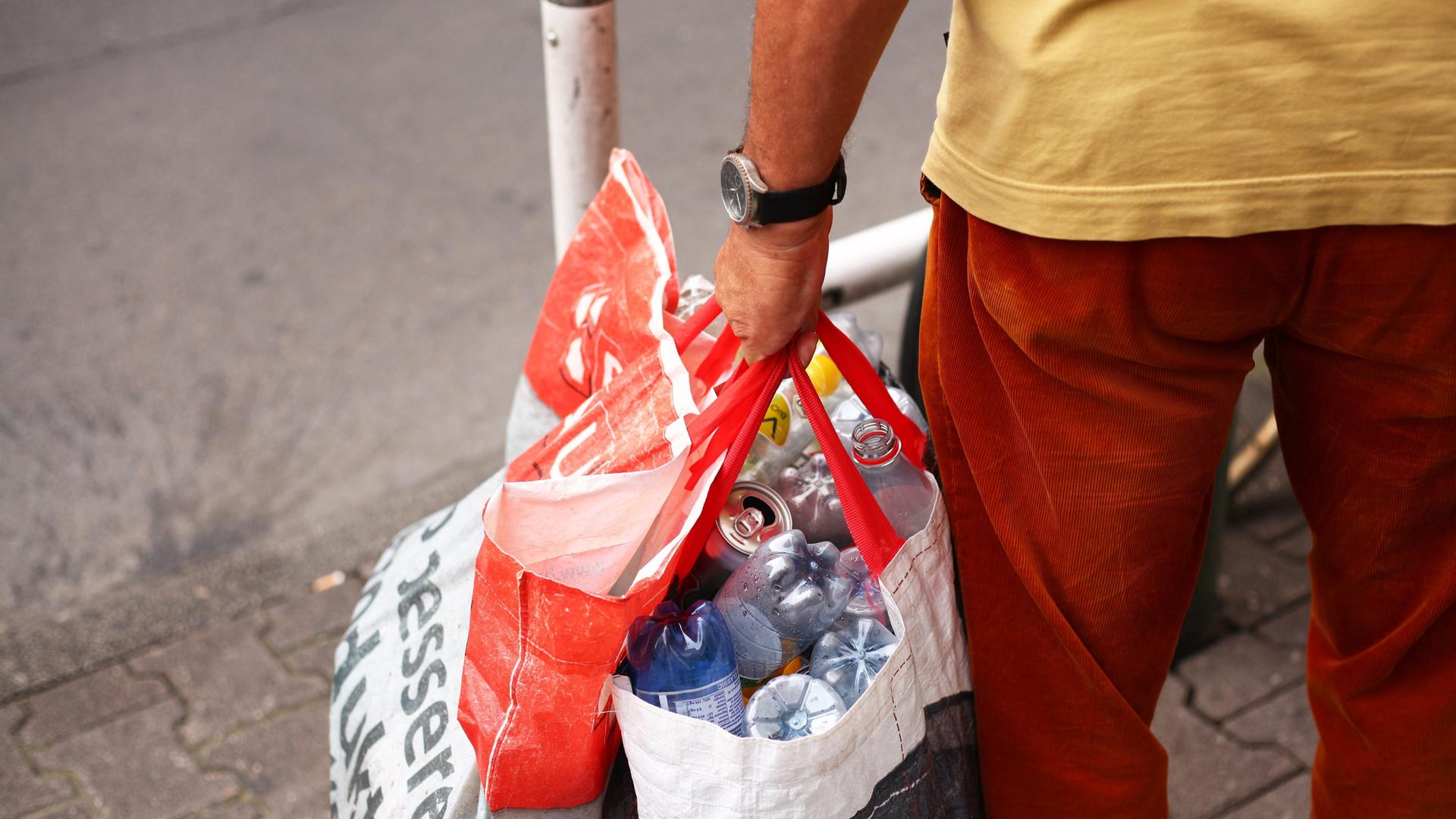 Detailaufnahme eines Mannes, der zwei Plastiktüten voller Pfandflaschen trägt.
