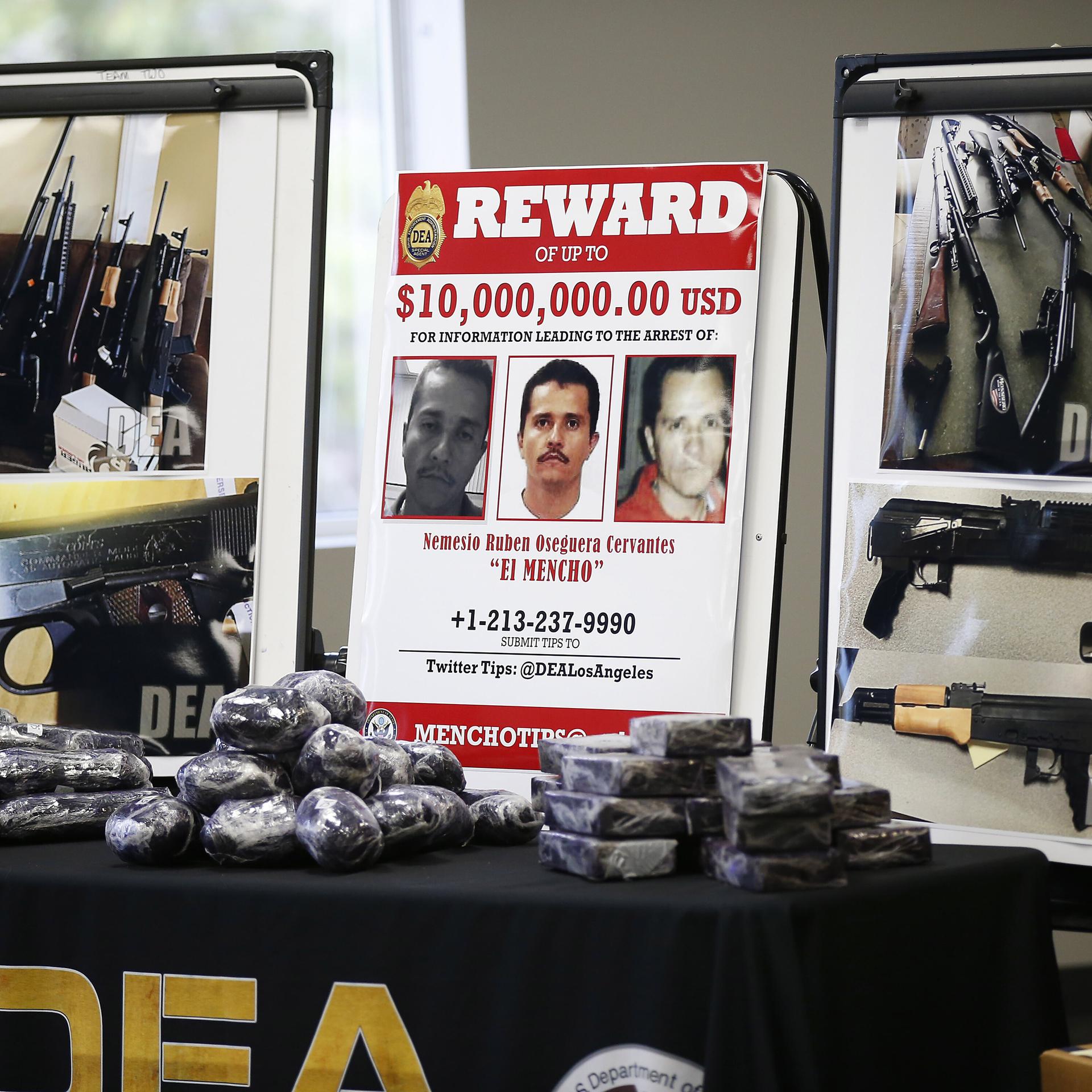 Es zeigt ein Fahndungsfoto des mexikanischen Drogenkartellboss Cervantes, auf den ein Kopfgeld von 10 Millionen US-Dollar ausgesetzt war. Daneben sind Fotos von Schusswaffen zu sehen. Vor den aufgestellten Bildern liegen schwarz-verpackte Päckchen.