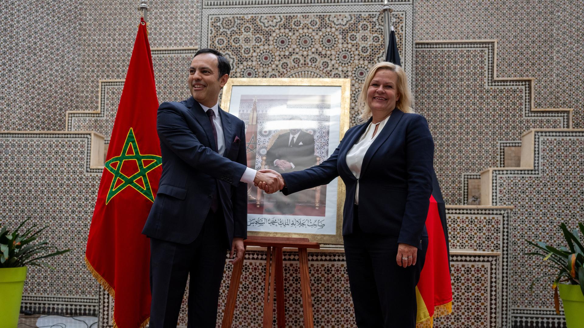 Younes Sekkouri schüttelt Nancy Faeser die Hand. Im Hintergrund steht ein Foto von König Mohammed VI zwischen Flaggen von Marokko und Deutschland.