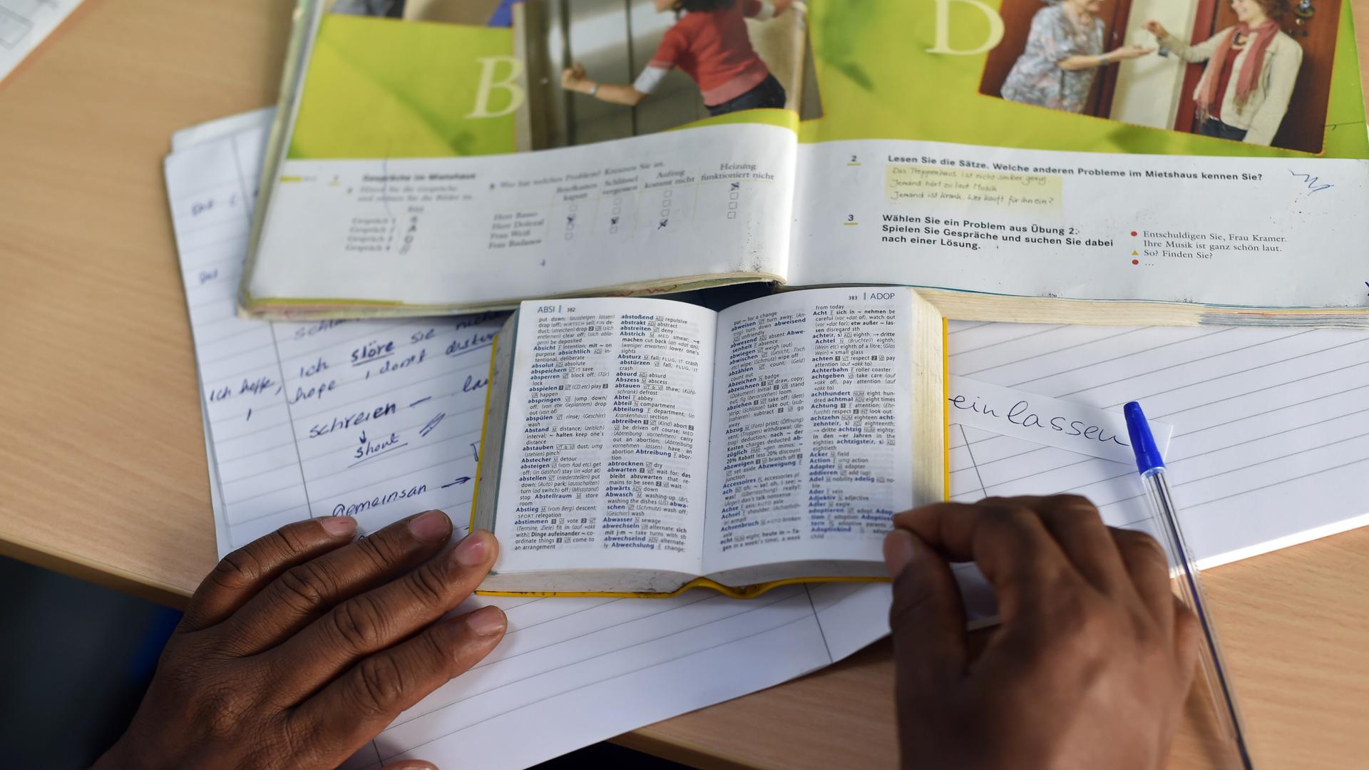 Eine Person nimmt an einem "Integrationskurs Deutsch" teil und hat vor sich Lehr- und Wörterbuch sowie ein Heft liegen.