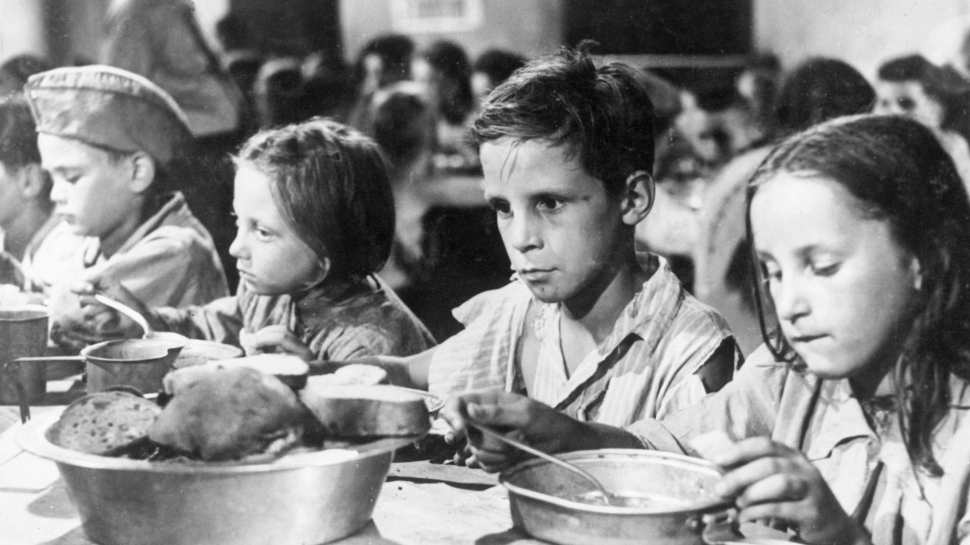 Mehrere Kinder essen auf dem Schwarz-Weiß-Bild an einem Tisch aus Blechtellern. Sie wirken ausgemergelt, die Augen eines Jungen sind leer.