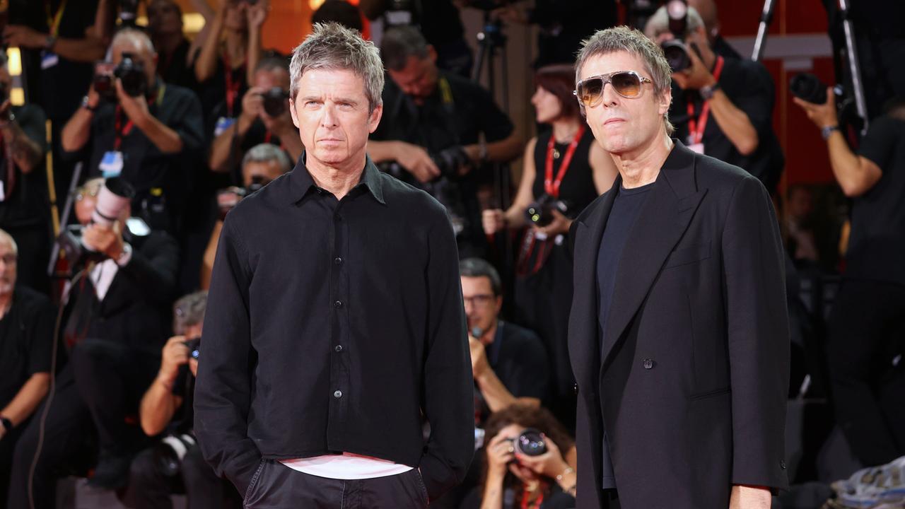 Noel Gallagher und Liam Gallagher stehen nebeneinander. Sie tragen schwarze Kleidung und gucken ein bisschen grimmig. Hinter ihnen sind viele Fotografen. Auch die Fotografen sind schwarz gekleidet. 