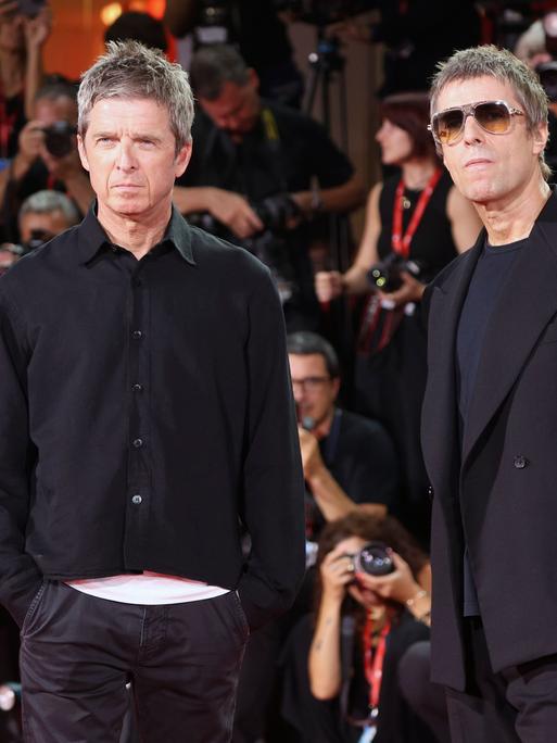 Noel Gallagher und Liam Gallagher stehen nebeneinander. Sie tragen schwarze Kleidung und gucken ein bisschen grimmig. Hinter ihnen sind viele Fotografen. Auch die Fotografen sind schwarz gekleidet. 