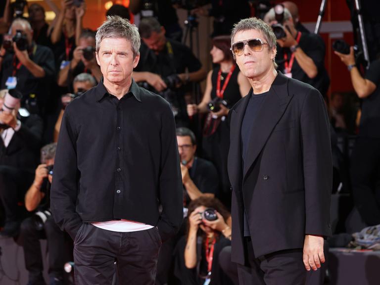 Noel Gallagher und Liam Gallagher stehen nebeneinander. Sie tragen schwarze Kleidung und gucken ein bisschen grimmig. Hinter ihnen sind viele Fotografen. Auch die Fotografen sind schwarz gekleidet. 