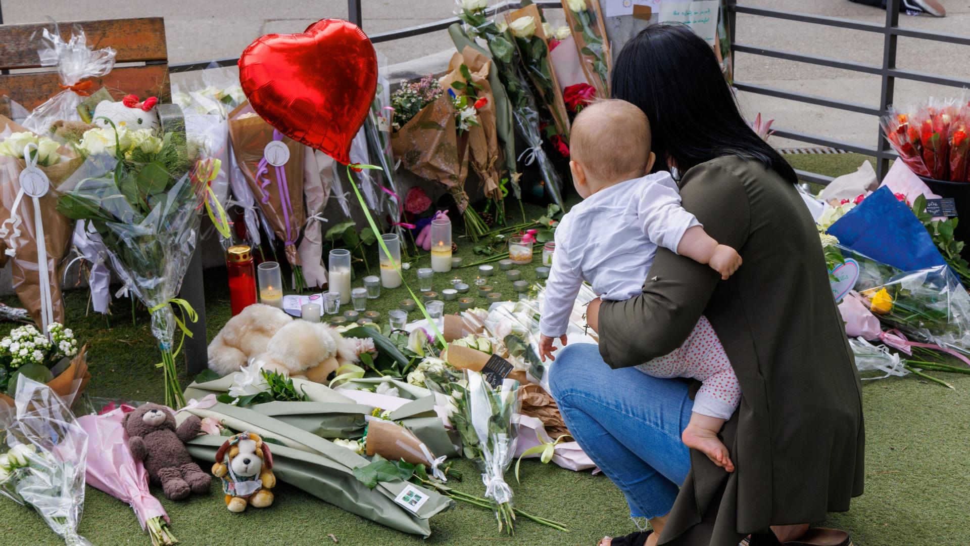 Eine Frau ist mit einem Kleinkind auf dem Arm in die Hocke gegangen und gedenkt der Opfer des Anschlags in der französischen Stadt Annecy. Auf dem BOden liegen Blumen, Stofftiere und ein LUftballon in Herzform. 