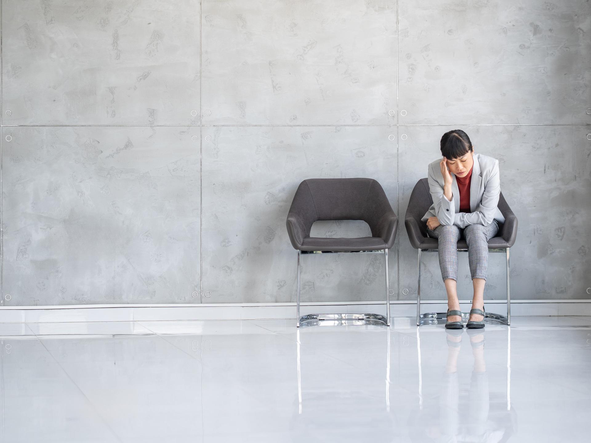 Eine Frau im grauen Business-Kostüm sitzt zusammengesunken auf einem Stuhl in einem Wartebereich.