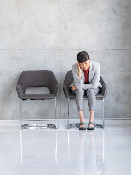 Eine Frau im grauen Business-Kostüm sitzt zusammengesunken auf einem Stuhl in einem Wartebereich.