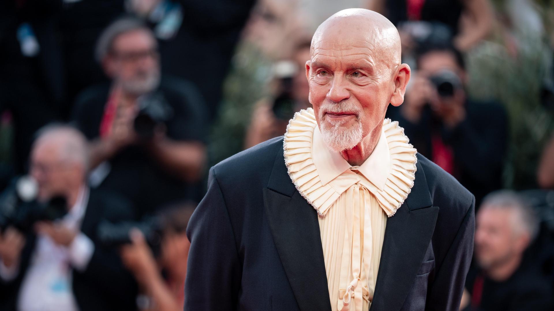 John Malkovich steht auf dem Roten Teppich und lässt sich fotografieren. Er trägt ein Hemd mit extravagantem Kragen.