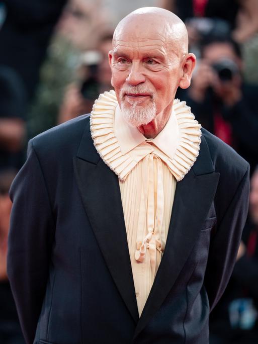 John Malkovich steht auf dem Roten Teppich und lässt sich fotografieren. Er trägt ein Hemd mit extravagantem Kragen.