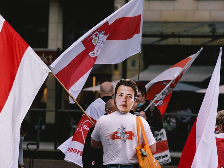 Verschiedene Menschen bei einer Demo mit den Flaggen von Belarus, einer trägt eine Maske von Roman Protassewitsch 