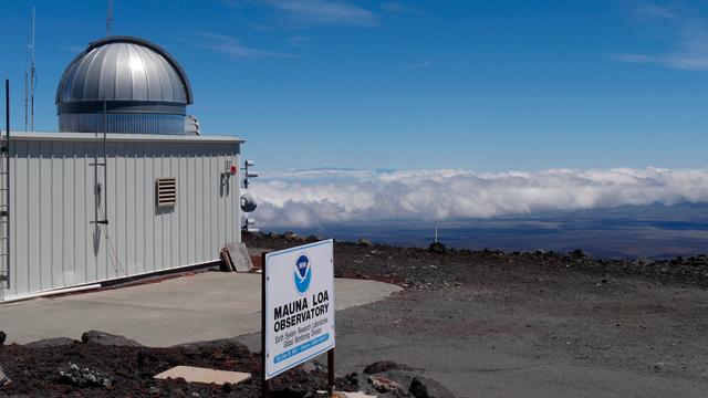 Ein flaches, weißes Messgebäude mit silberner Beobachtungskuppel auf einer kargen, dunklen Lavaebene unter blauem Himmel. Im Vordergrund ein Schild mit der Aufschrift „Mauna Loa Observatory". Ein flaches, weißes Messgebäude mit silberner Beobachtungskuppel auf einer kargen, dunklen Lavaebene unter blauem Himmel. Im Vordergrund ein Schild mit der Aufschrift „Mauna Loa Observatory".