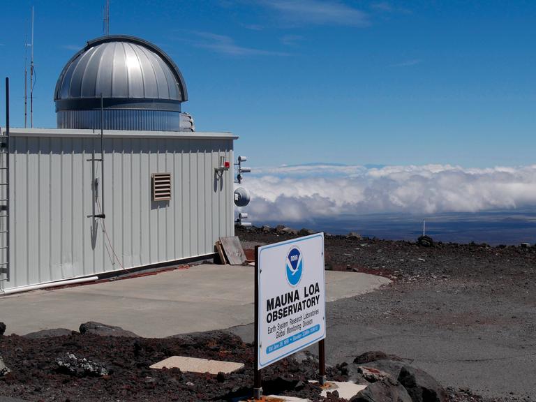 Ein flaches, weißes Messgebäude mit silberner Beobachtungskuppel auf einer kargen, dunklen Lavaebene unter blauem Himmel. Im Vordergrund ein Schild mit der Aufschrift „Mauna Loa Observatory". Ein flaches, weißes Messgebäude mit silberner Beobachtungskuppel auf einer kargen, dunklen Lavaebene unter blauem Himmel. Im Vordergrund ein Schild mit der Aufschrift „Mauna Loa Observatory".