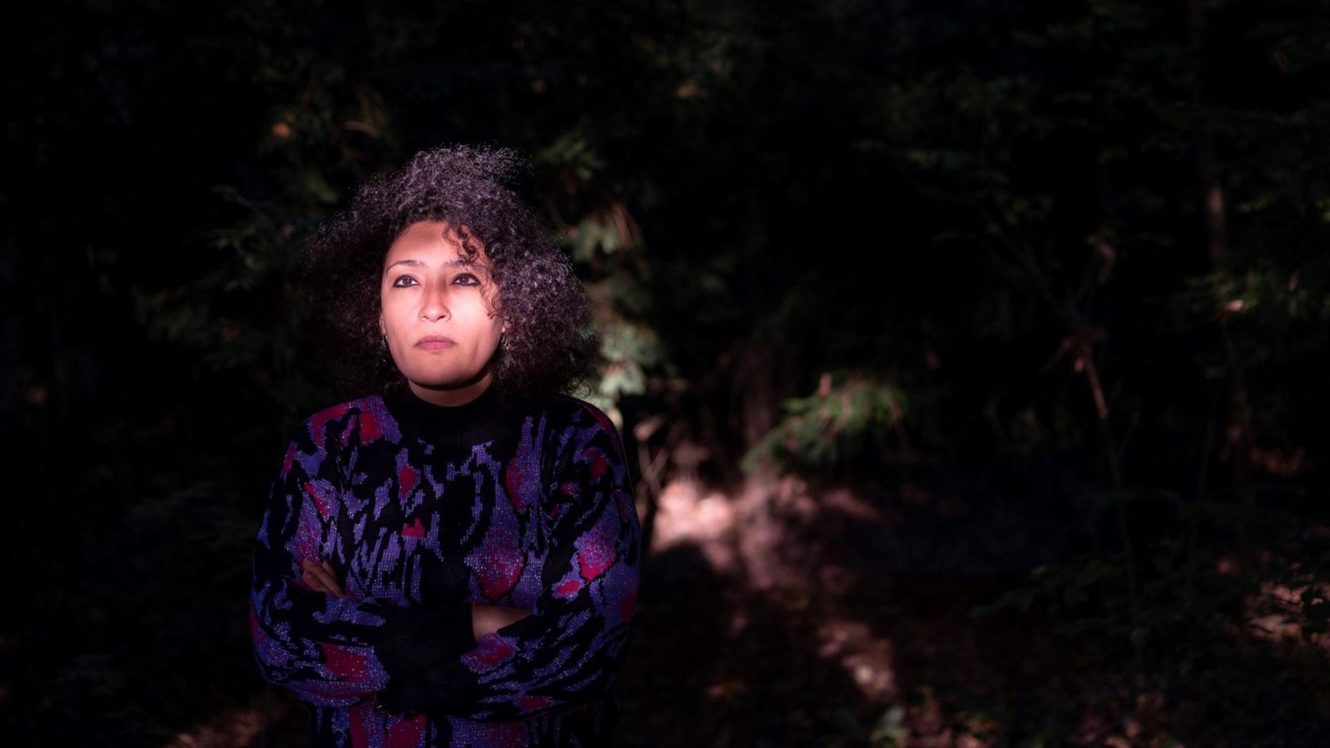 Eine junge Frau sitzt in einem dunklen Gartenareal, während ihr Gesicht hell erleuchtet wird.