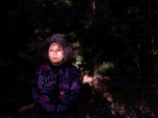 Eine junge Frau sitzt in einem dunklen Gartenareal, während ihr Gesicht hell erleuchtet wird.