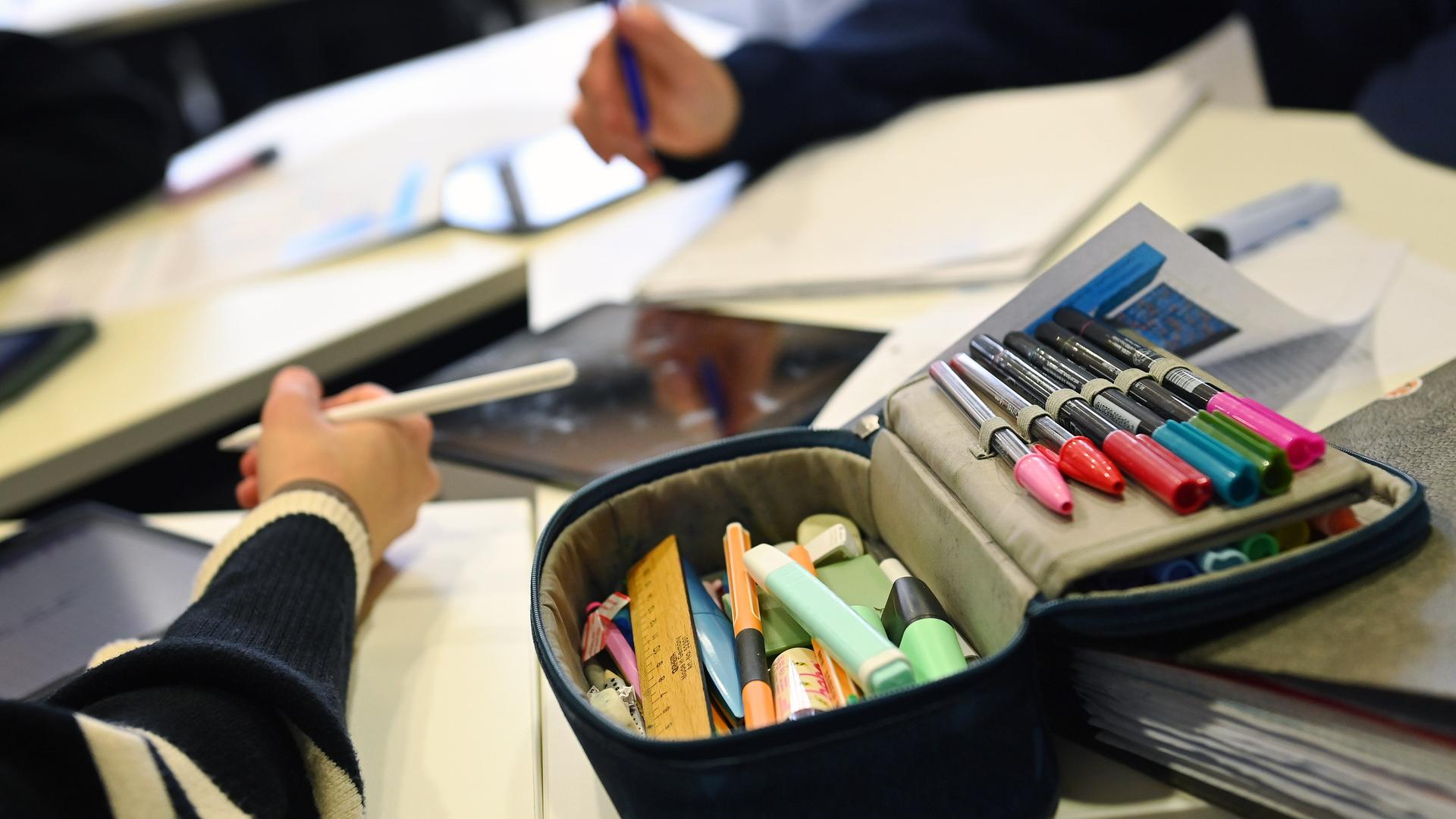 Schulunterricht in Bayern: Auf dem Tisch liegt ein Mäppchen mit bunten Filzstiften, zwei Kinderarme halten Stifte in der Hand.