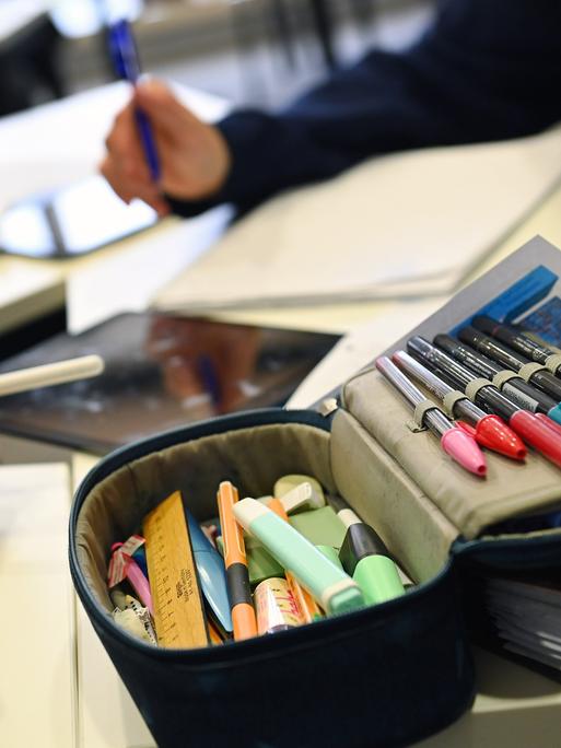 Schulunterricht in Bayern: Auf dem Tisch liegt ein Mäppchen mit bunten Filzstiften, zwei Kinderarme halten Stifte in der Hand.