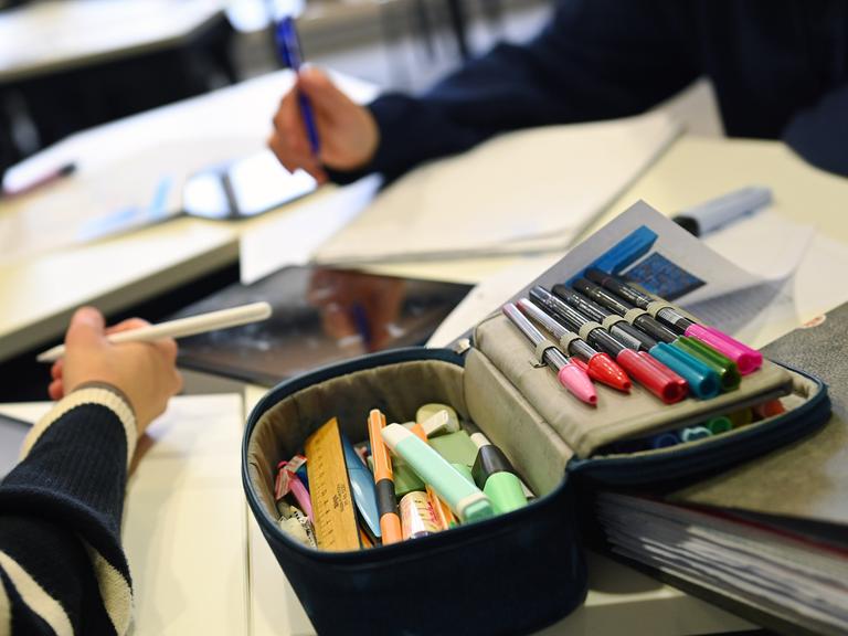 Schulunterricht in Bayern: Auf dem Tisch liegt ein Mäppchen mit bunten Filzstiften, zwei Kinderarme halten Stifte in der Hand.