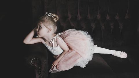 Ein Mädchen liegt in einem Tutu, den Kopf aufgestützt, auf einem dunkelbraunen Sofa