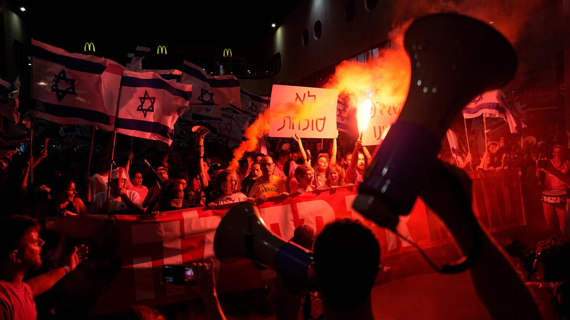 Protestteilnehmer im Schein von Pyrotechnik halten Plakate, israelische Fahnen und Megafone.