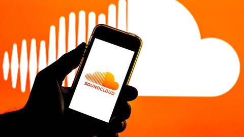In der Illustration ist eine Hand zu sehen, die ein Smartphone hält. Auf dem Bildschirm ist das Soundcloud-Logo, auch im Hintergrund ist das Logo, eine weiße Wolke auf orangem Grund, wobei die Wolke links gestrichelt ist.