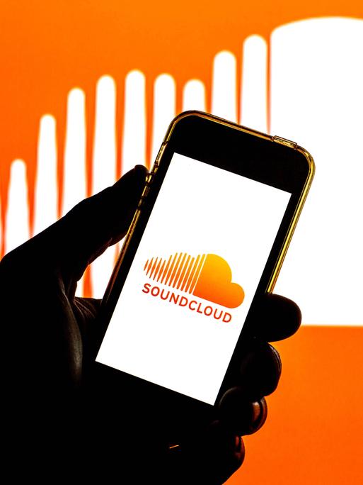 In der Illustration ist eine Hand zu sehen, die ein Smartphone hält. Auf dem Bildschirm ist das Soundcloud-Logo, auch im Hintergrund ist das Logo, eine weiße Wolke auf orangem Grund, wobei die Wolke links gestrichelt ist.