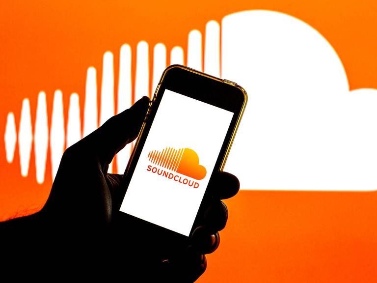In der Illustration ist eine Hand zu sehen, die ein Smartphone hält. Auf dem Bildschirm ist das Soundcloud-Logo, auch im Hintergrund ist das Logo, eine weiße Wolke auf orangem Grund, wobei die Wolke links gestrichelt ist.