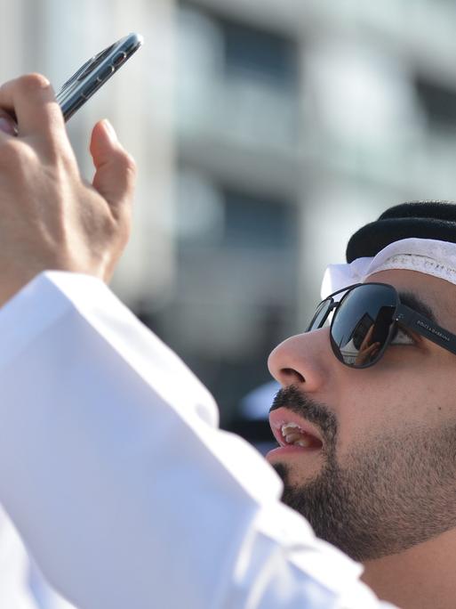 Scheich Mansour Bin Mohammed Bin Rashid Al Maktoum an der Ziellinie der Dubai Meraas Stage in Dubai, mit seinem Smartphone das er in die Höhe hält.
