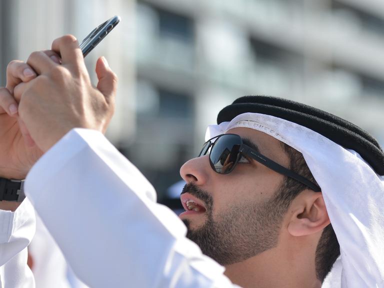 Scheich Mansour Bin Mohammed Bin Rashid Al Maktoum an der Ziellinie der Dubai Meraas Stage in Dubai, mit seinem Smartphone das er in die Höhe hält.