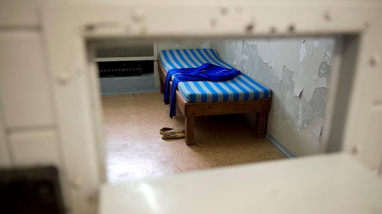 Durch eine Türöffnung ist eine Zelle zu sehen, in der ein Bett mit blau gestreifter Matratze steht. Obenauf liegt eine Hose, davor stehen Pantoffeln auf dem Boden; der Putz an der Wand ist großflächig abgeblättert.