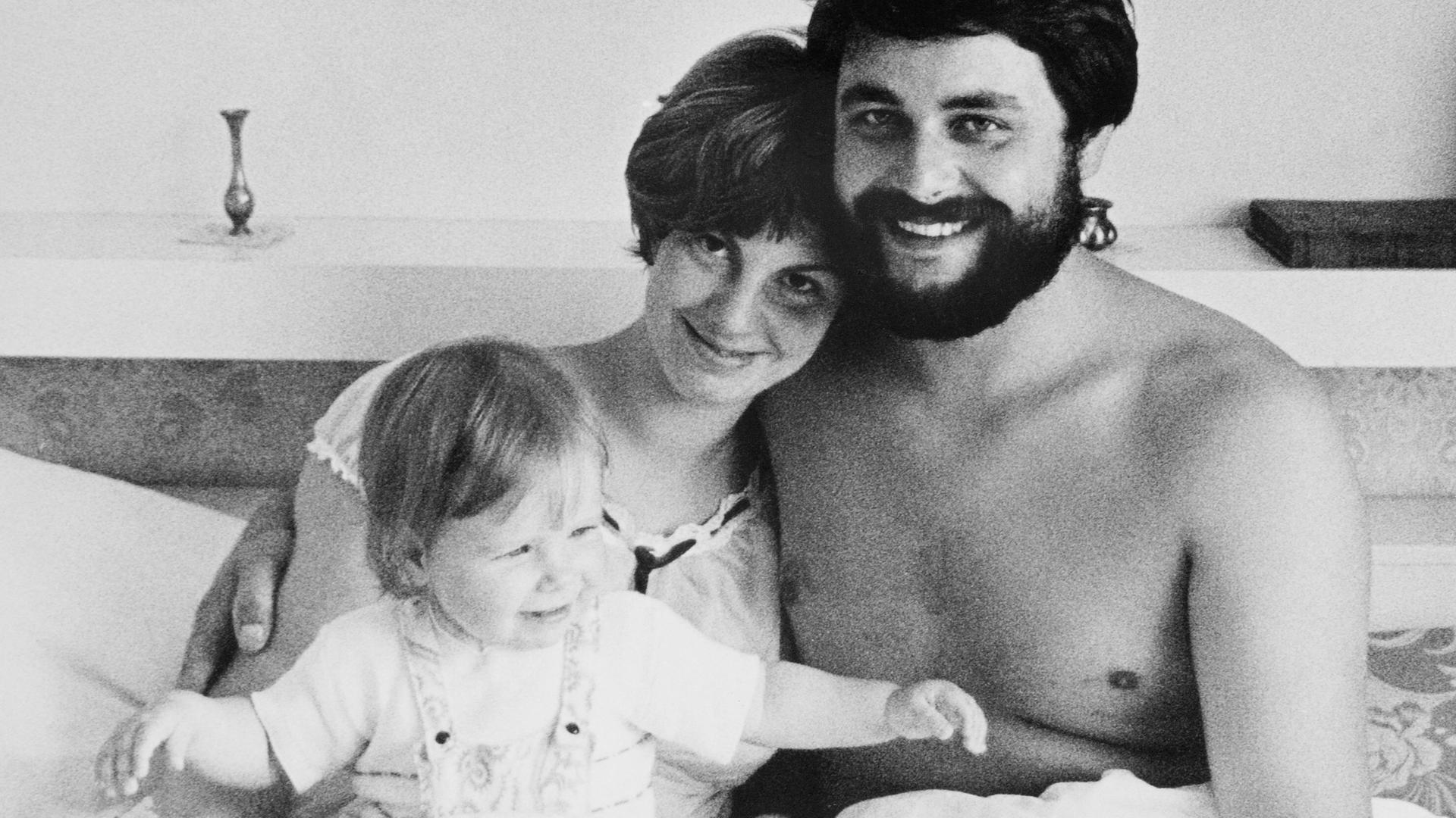 Schwarzweiss Aufnahme von einem jungen Paar mit Baby im Bett, die Eltern lächeln glücklich in die Kamera. DDR, Leipzig 1978.