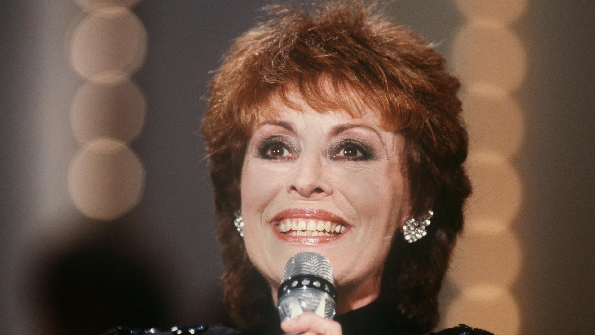 Caterina Valente im April 1987. Sie hat braunes Haar und trägt große silberne Ohrringe, in der Hand hält sie ein Mikrofon. 