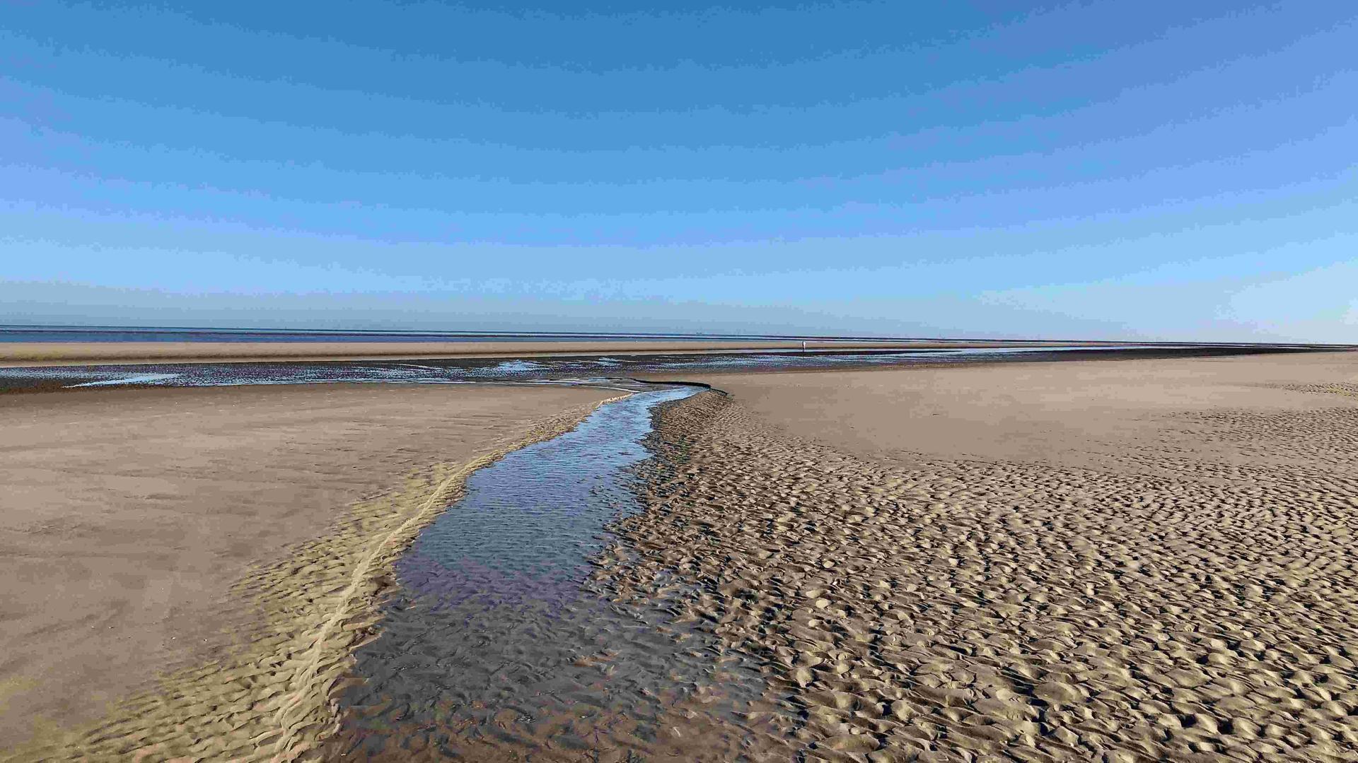 Ein Bächlein im Watt und der Ausblick aufs Wattenmeer unter blauem Himmel.