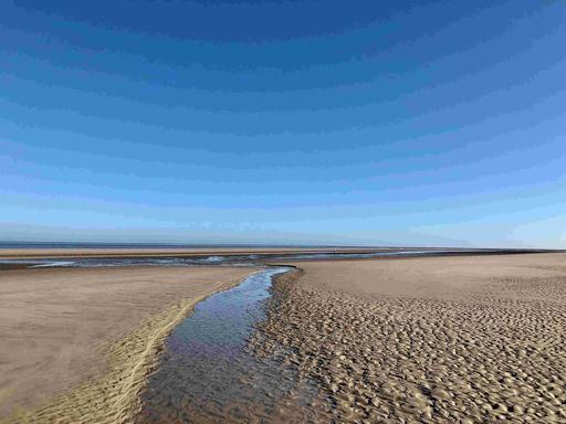 Ein Bächlein im Watt und der Ausblick aufs Wattenmeer unter blauem Himmel.