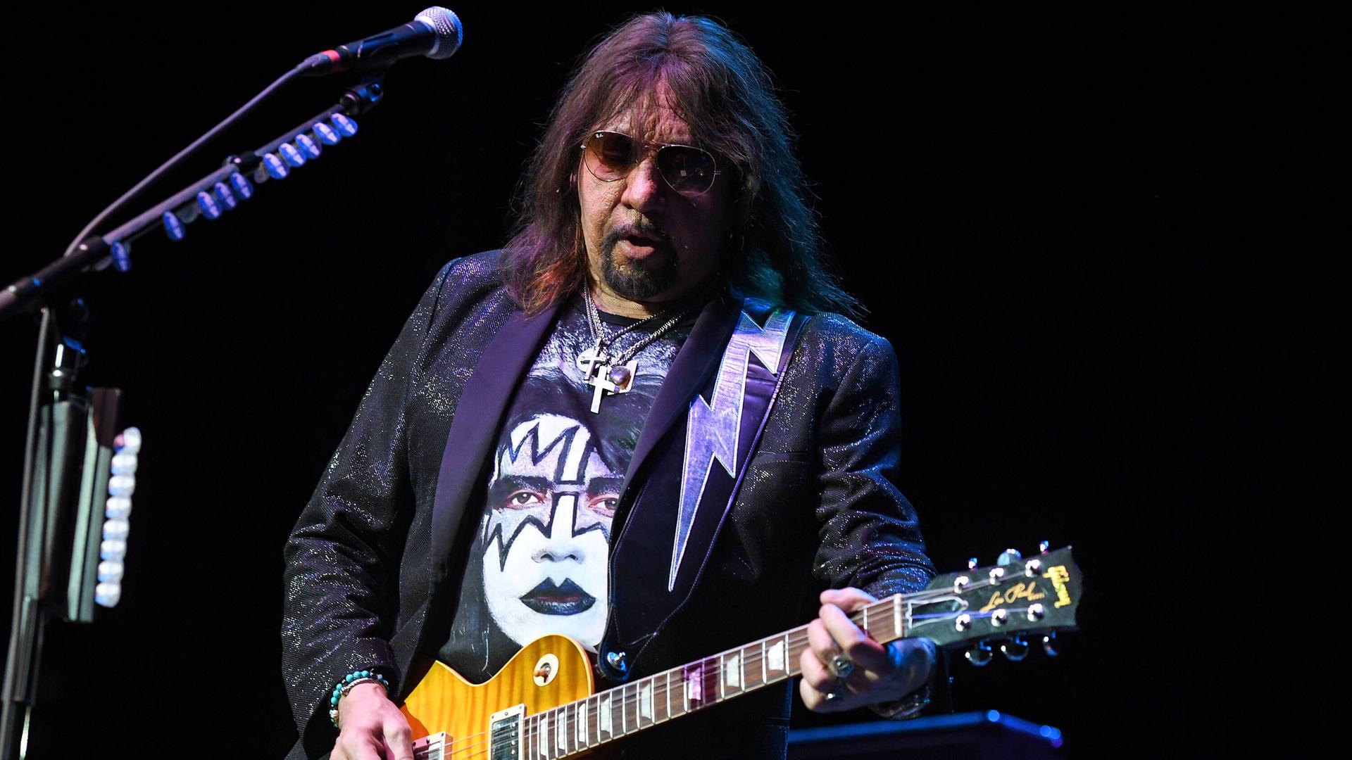 Das Bild zeigt Ace Frehley mit einer Sonnenbrille beim Spiel seiner E-Gitarre auf einer Bühne. Auf seinem T-Shirt ist eine Abbildung seines Gesichts mit dem typischen schwarz-weißen Make-Up aus Zeiten der Band "Kiss" zu sehen. Das Bild zeigt Ace Frehley mit einer Sonnenbrille beim Spiel seiner E-Gitarre auf einer Bühne. Auf seinem T-Shirt ist eine Abbildung seines Gesichts mit dem typischen schwarz-weißen Make-Up aus Zeiten der Band "Kiss" zu sehen.