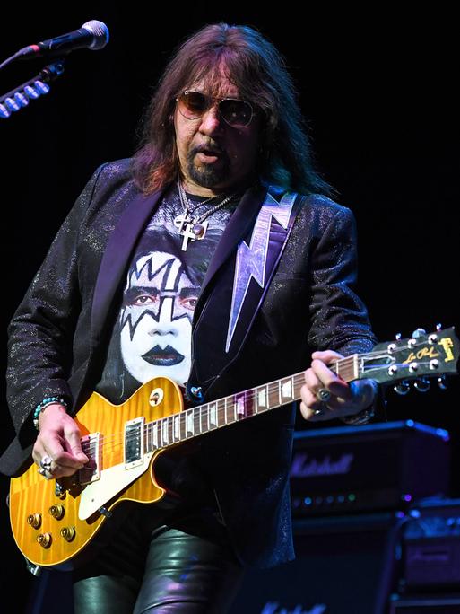Das Bild zeigt Ace Frehley mit einer Sonnenbrille beim Spiel seiner E-Gitarre auf einer Bühne. Auf seinem T-Shirt ist eine Abbildung seines Gesichts mit dem typischen schwarz-weißen Make-Up aus Zeiten der Band "Kiss" zu sehen.