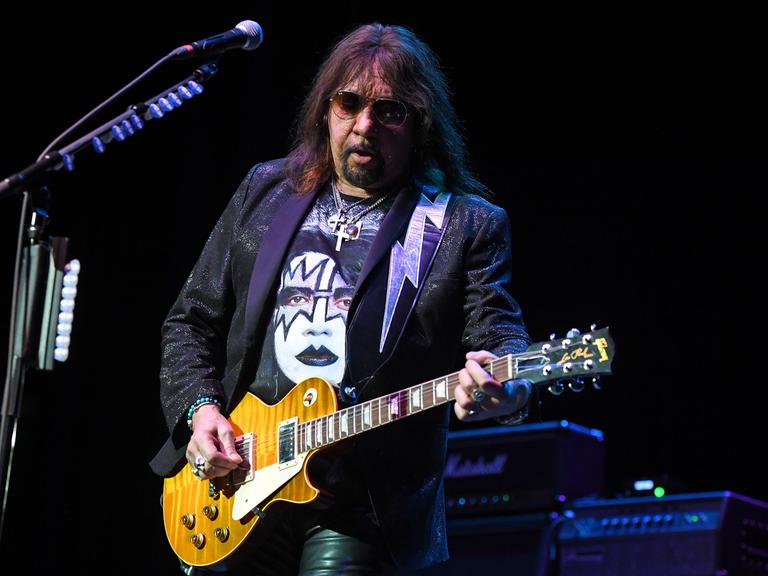 Das Bild zeigt Ace Frehley mit einer Sonnenbrille beim Spiel seiner E-Gitarre auf einer Bühne. Auf seinem T-Shirt ist eine Abbildung seines Gesichts mit dem typischen schwarz-weißen Make-Up aus Zeiten der Band "Kiss" zu sehen.