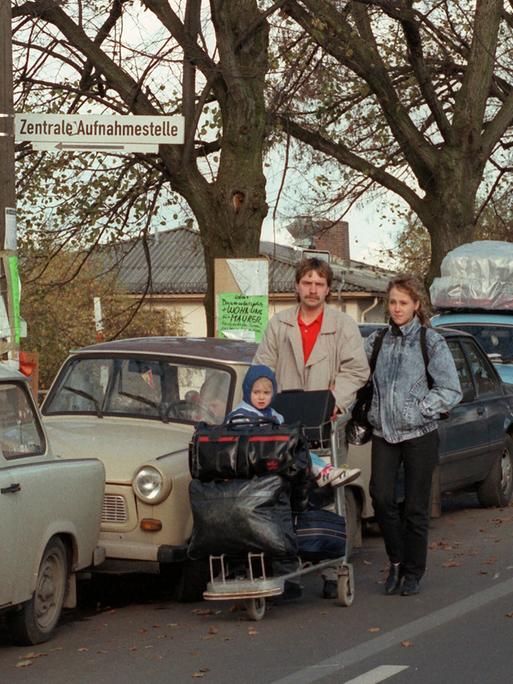 Eine Familie läuft an geparkten Trabanten vorbei. Ihr Kind sitzt auf einem Plattformwagen, der mit Koffern und Taschen voll bepackt ist. Eine Familie läuft an geparkten Trabanten vorbei. Ihr Kind sitzt auf einem Plattformwagen, der mit Koffern und Taschen voll bepackt ist.