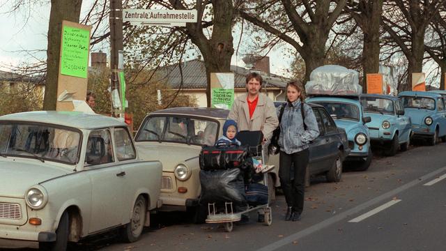 Eine Familie läuft an geparkten Trabanten vorbei. Ihr Kind sitzt auf einem Plattformwagen, der mit Koffern und Taschen voll bepackt ist.