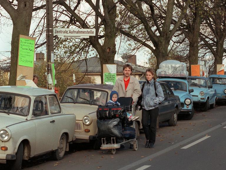Eine Familie läuft an geparkten Trabanten vorbei. Ihr Kind sitzt auf einem Plattformwagen, der mit Koffern und Taschen voll bepackt ist. Eine Familie läuft an geparkten Trabanten vorbei. Ihr Kind sitzt auf einem Plattformwagen, der mit Koffern und Taschen voll bepackt ist.